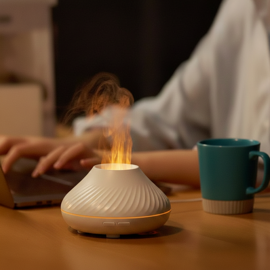 Black essential oil diffuser with flames on a wooden table, person using laptop in the background.